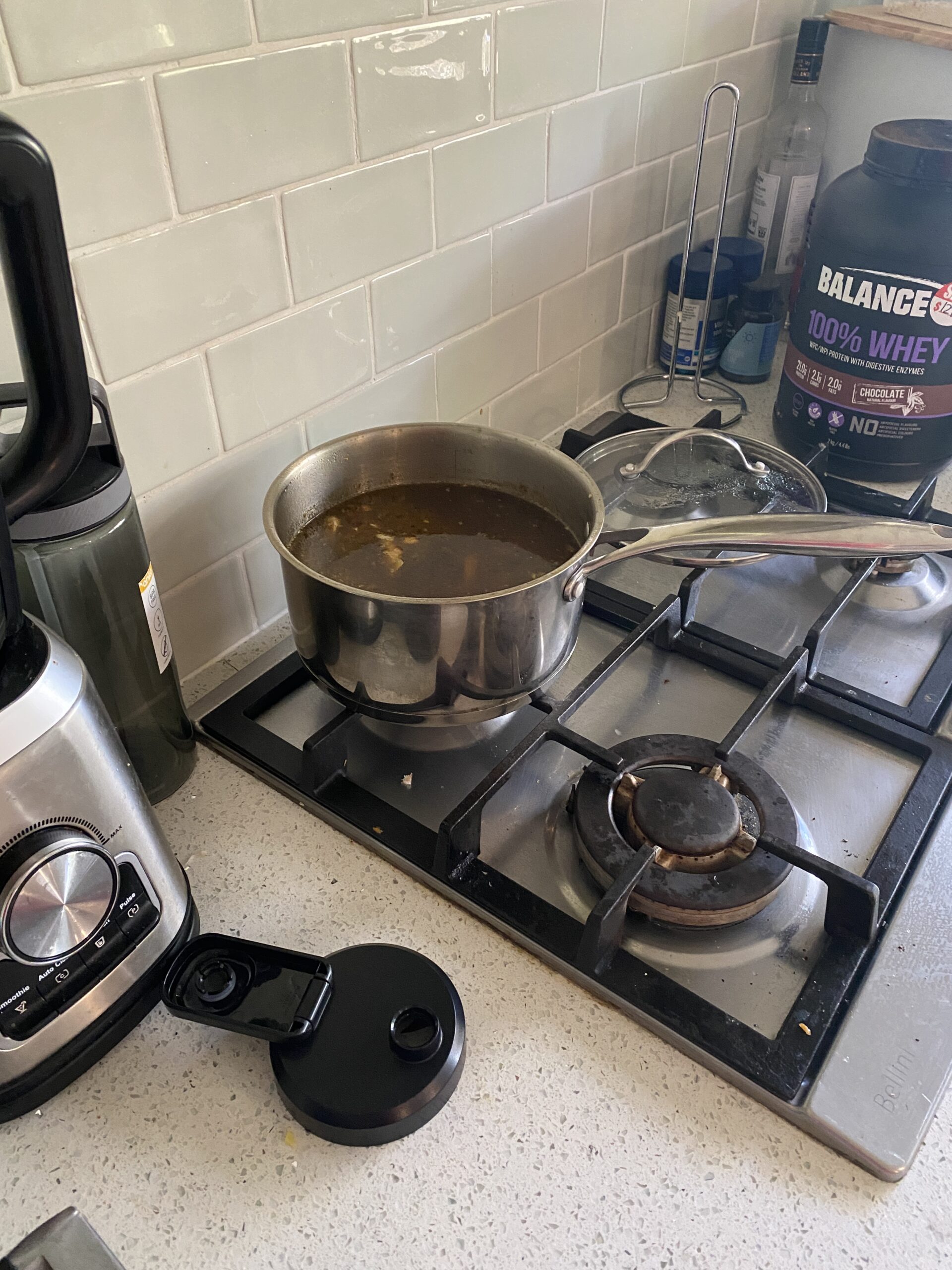 a stove with a pot of chicken broth and soup top - there are gas burners on the left of the image, which are turned OFF, there is also a top lid of a protein shaker on the far left