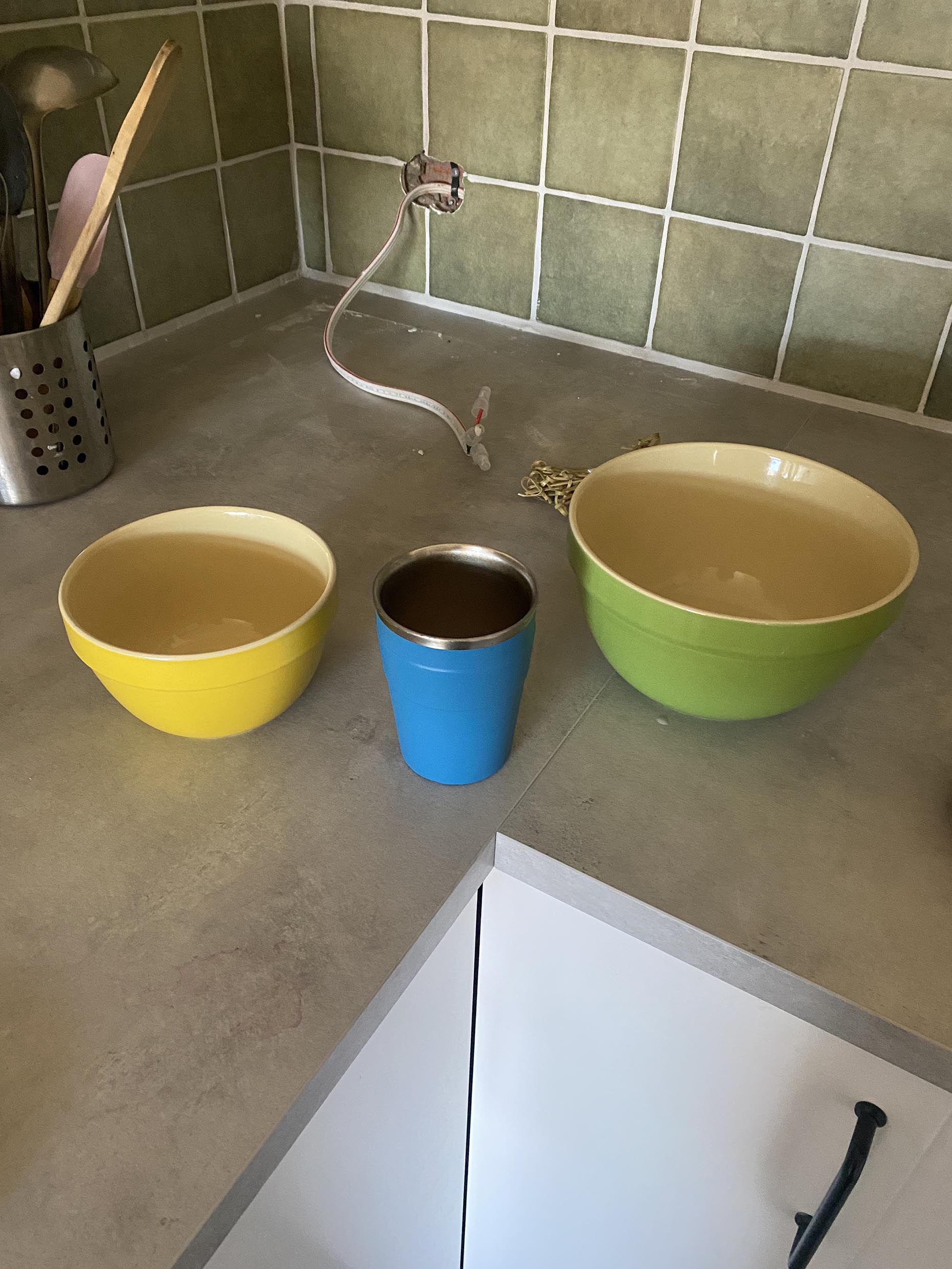 image shows three objects on a concrete coloured bench top with green tiles, moon white grout and electrical works behind. The three bowls are a yellow small bowl, a medium blue keep cup and a larger green mixing bowl (ceramic)