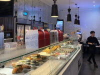glass cabinet with sandwiches and cakes and cake boxes on top, a waiter walking to the right of the image and black metal lights hanging from the ceiling