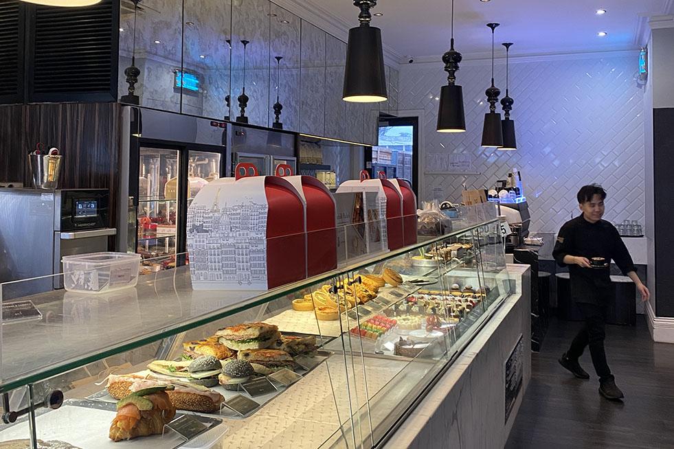 glass cabinet with sandwiches and cakes and cake boxes on top, a waiter walking to the right of the image and black metal lights hanging from the ceiling