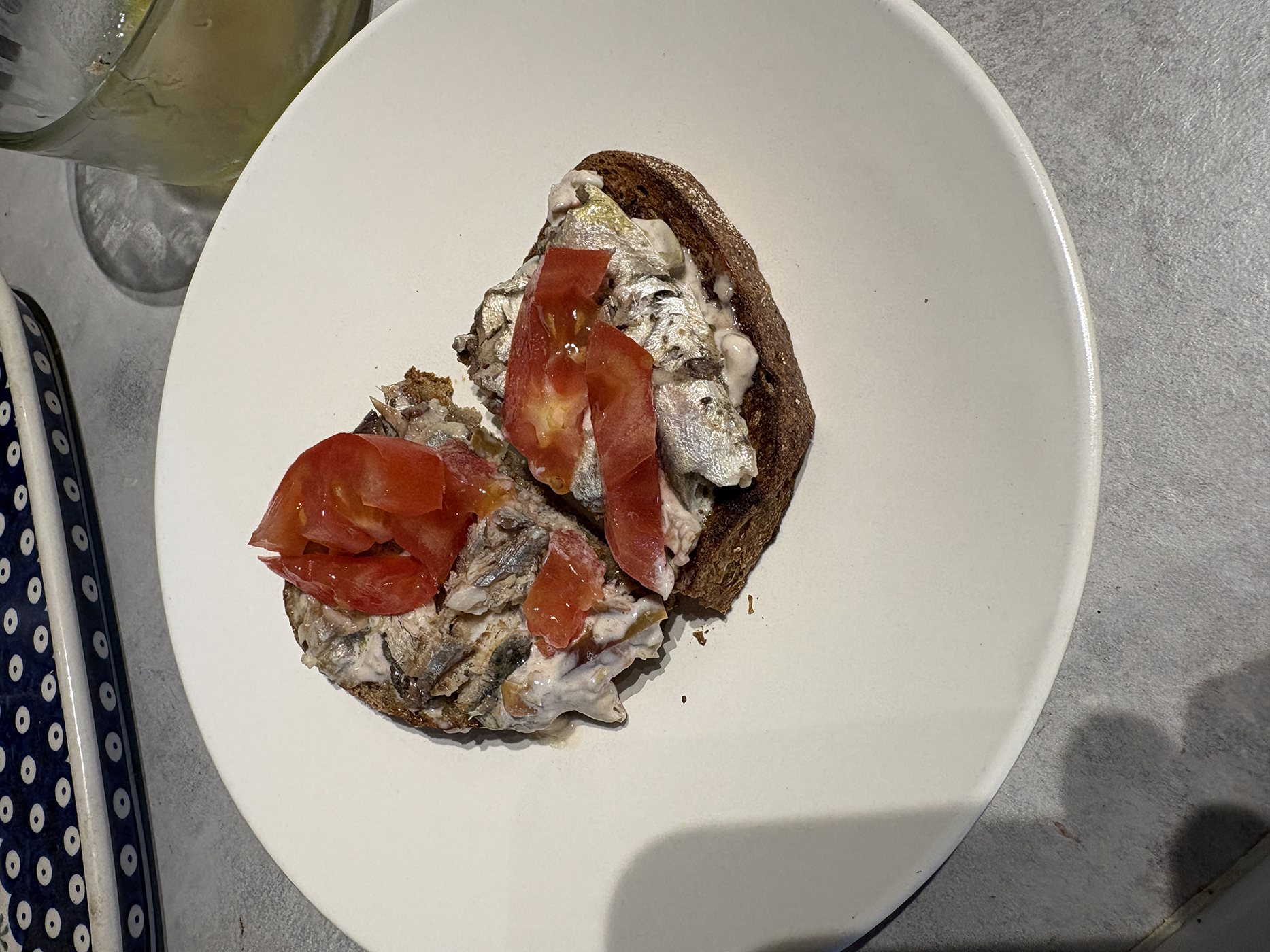 a white small plate with a piece of toast topped with olive dip, sardines and slices of tomato